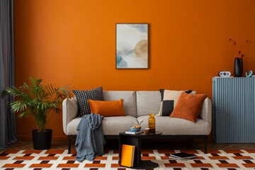 Living room with a bright orange wall, light grey sofa, patterned cushions, and a geometric rug,...