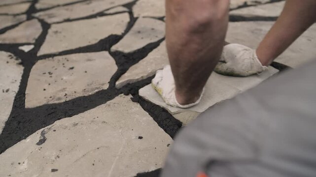 Worker hands in gloves setting natural flagstone into dark mortar to build a garden patio or pathway, close-up detail of masonry work