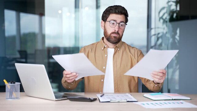 Confused businessman having difficulty with paperwork sitting at desk at workplace in modern business office. Frustrated financier in glasses unhappy with financial results browsing reading documents