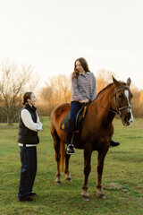 Fototapeta premium A young girl sits confidently on a horse while a smiling friend stands beside her. The scene captures a tranquil outdoor setting during golden hour, with soft light filtering through trees.
