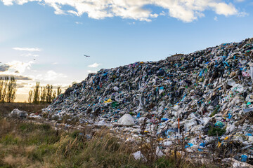 A large landfill contains heaps of garbage and waste materials under cloudy skies with some birds flying above the site