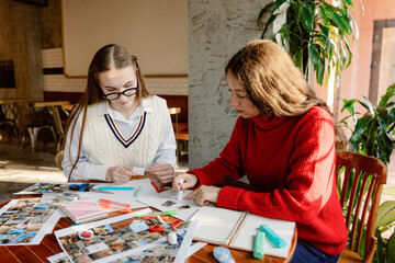 Two friends enjoy a creative afternoon in a warm cafe, working on collages with scissors, glue, and colorful markers while surrounded by plants and cheerful decor.