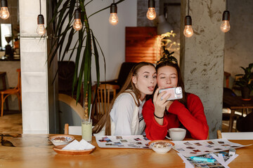 Two young women share a joyful moment in a cozy cafe, surrounded by green plants and warm lighting. They capture memories while sipping drinks and smiling widely, creating a carefree atmosphere.