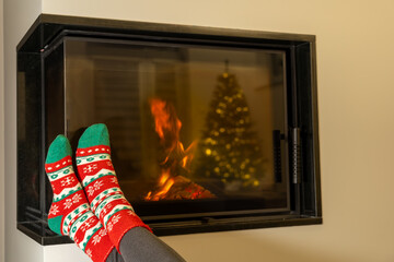 Women's feet in thick socks warming at fireplace. The fire and the Christmas tree are visible in the glass. Minimalist, modern interior