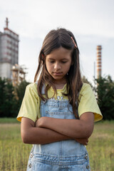 Thoughtful girl standing in front of industrial chimneys with arms crossed and serious expression. The portrait symbolizes climate anxiety, future concerns and environmental responsibility.