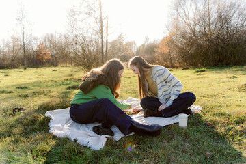 Fototapeta premium Two friends sit on a blanket in a vibrant green field, surrounded by trees. They engage in a creative activity, laughing and sharing moments under a clear sky during a pleasant afternoon.