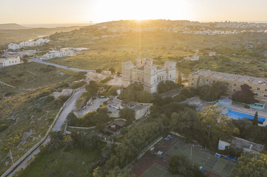 Aerial view of Castello Zammitello bathed in the golden light of dawn, amidst the undulating landscape and azure pool contrasting with the lush greenery, Mistra bay, St. Paul's Bay, Malta.