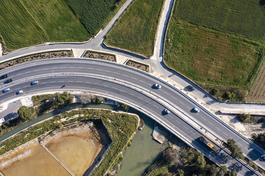 Aerial view of a winding highway cutting through vibrant green fields and brown patches, with cars traversing the asphalt ribbon, Bugibba, St. Paul's Bay, Malta.