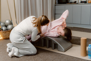 In a serene exercise studio, a trainer assists a young girl in perfecting a stretching pose on an incline. The space is equipped with workout tools, promoting wellness and fitness.