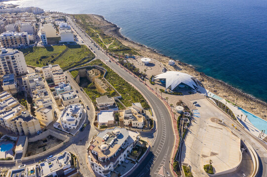 Aerial view of buildings and roads meet the rugged coastline, contrasting urban development with the natural sea, Bugibba, St. Paul's Bay, Malta.