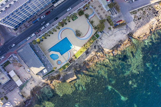 Aerial view of turquoise waters meet the rocky coast beside a hotel with a pool and palm trees, Bugibba, St. Paul's Bay, Malta.