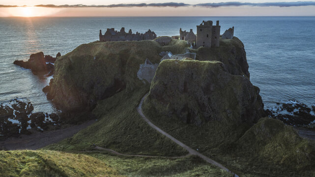 Aerial view of Dunnottar Castle ruins perched dramatically on a cliffside, where golden sunlight kisses the rugged coastline, Stonehaven, Scotland, United Kingdom.