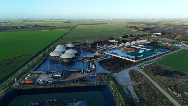 Aerial view of biogas plant with rounded tanks and adjacent farm buildings amidst expansive green fields under a muted sky, Wisbech, England, United Kingdom.