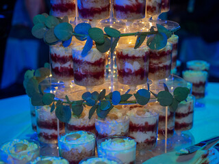 multi-tiered display of individual dessert trifles featuring layers of white cream, sponge cake, and red berry compote. The tower is decorated with fresh eucalyptus leaves and warm fairy lights
