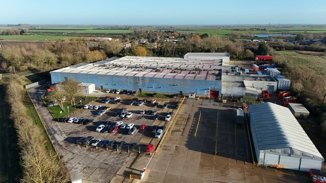 Aerial view of a large industrial building with a vast parking lot filled with cars under a soft winter sky, Ely, England, United Kingdom.