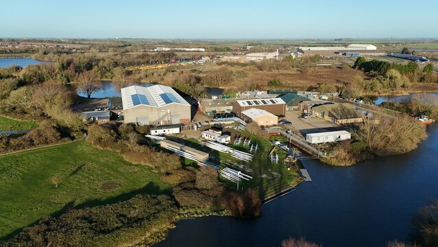 Aerial view of industrial buildings and boats nestled beside the tranquil, dark waters of the river, a stark contrast to the bright green grass, Ely, England, United Kingdom.