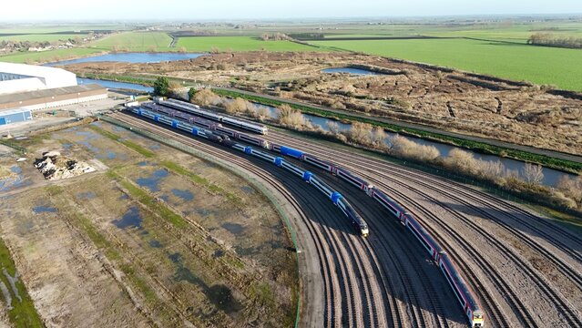 Aerial view of trains standing in formation near silvery water canals and green fields under a clear sky, Ely, England, United Kingdom.