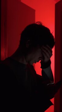 Man shocked by bad news on smartphone, standing in doorway under red light, reading negative headlines alone at home. Vertical video