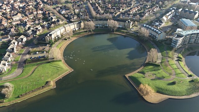 Aerial view of a serene, circular lake reflecting the pale winter sky, bordered by curved walkways and modern buildings, a tranquil oasis, Milton Keynes, England, United Kingdom.
