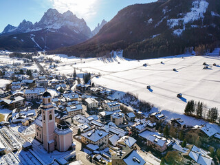 Aerial view of a charming village nestled in a snow-blanketed valley, framed by the majestic Dolomites under a clear sky, Sexten, Trentino-South Tyrol, Italy.
