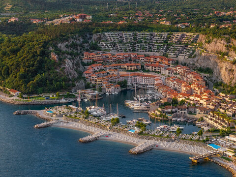 Aerial view of the marina's turquoise waters meet the terracotta rooftops and creamy stone buildings nestled against the verdant hillside, Portopiccolo Sistiana, Friuli-Venezia Giulia, Italy.