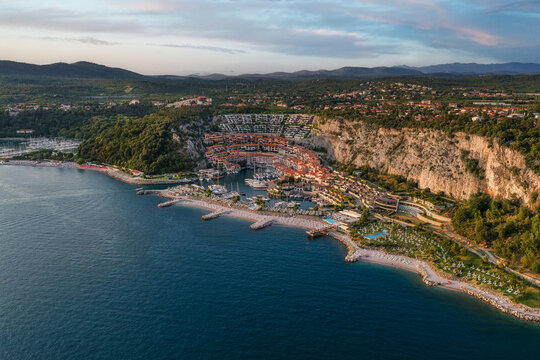 Aerial view of a coastal town nestled beside the glistening sea, where vibrant buildings contrast with the rugged cliffs and lush greenery, Portopiccolo Sistiana, Friuli-Venezia Giulia, Italy.