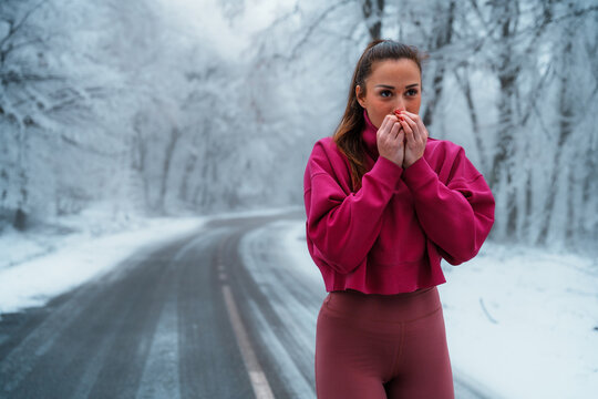 Young woman standing on a snowy forest road in winter, warming her hands to protect from cold while taking a break from her active outdoor fitness workout - Powered by Adobe