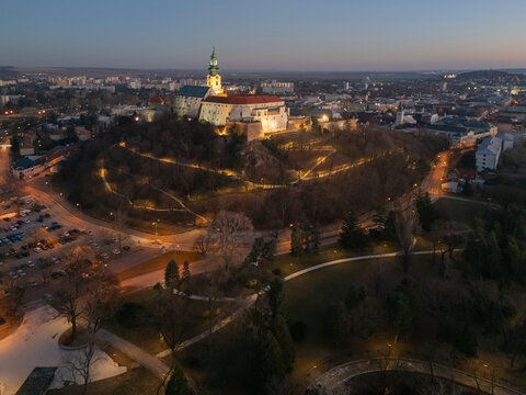 Aerial view of Nitra Castle perched atop a hill, illuminated pathways wind through the dark trees below, contrasting with the cityscape in the distance, Nitra, Nitriansky kraj, Slovakia.