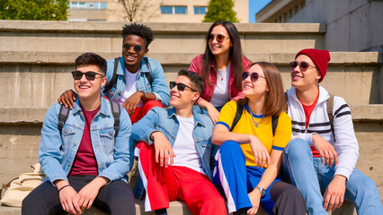 Six young people (boys and girls) of different nationalities sit on concrete steps on a sunny day. The students are dressed in bright casual clothes, many wearing sunglasses. They smile and look in th