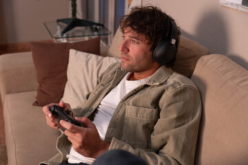 Young man engrossed in playing video games on a sofa, wearing headphones and holding a controller, finding relaxation and entertainment during his free time