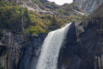 Bridalveil Fall water powerfully cascading down the ancient granite cliff in Yosemite National Park, surrounded by lush green foliage and rocky mountain slopes