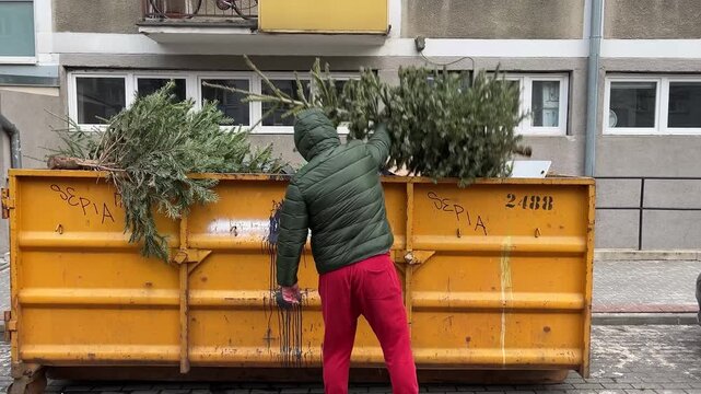 Man in a hooded jacket throwing a used christmas tree into a large yellow dumpster on the street after the holiday season, symbolizing the end of festivities and proper waste disposal