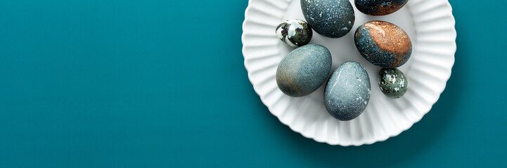 A collection of intricately decorated Easter eggs in various shades rests on a white plate against a soothing teal backdrop, perfect for spring festivities and family traditions, banner