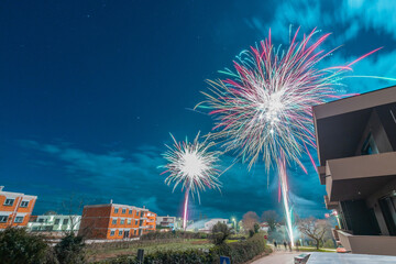 Colorful fireworks burst across a deep blue night sky above a quiet residential neighborhood, illuminating modern buildings and trees, capturing a festive celebration with dynamic light trails and