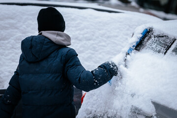 Individual wearing a dark winter jacket is clearing snow from a car windshield with a blue brush, showcasing winter weather challenges and personal effort in snow removal