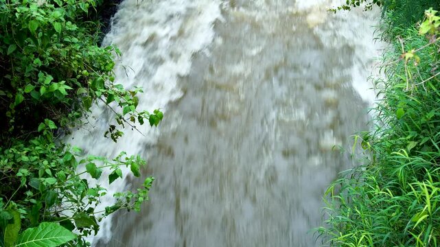 Rapid muddy water flowing strongly through a narrow ditch lined with green grass. High angle view of turbulent brown river current rushing between vegetation banks during flood or storm aftermath.