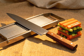 A composition of a knife, a steel mandoline, and a stack of identical carrot and cucumber sticks on textured linen canvas