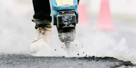Construction worker using a jackhammer breaking asphalt road surface for renovation, creating dust and debris