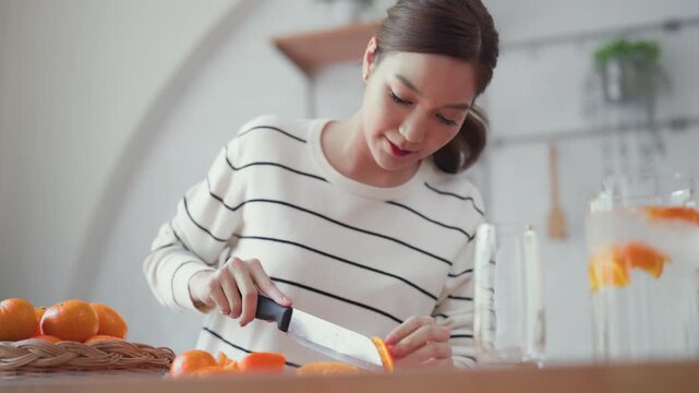 Beautiful young asian woman slicing a fresh orange on a wooden board in a kitchen, focusing on healthy diet and nutrition for longevity and wellness.