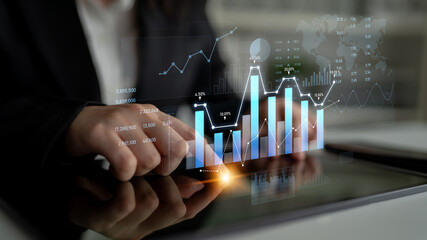 A business professional works on a tablet, analyzing digital graphs and charts that illustrate...