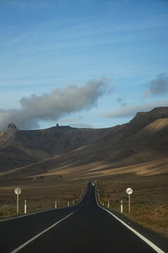 Perspectiva desde el asfalto de una carretera recta que se dirige hacia las monta&ntilde;as escarpadas del macizo de Famara bajo un cielo nublado, capturando la esencia de un viaje por carretera en Lanzarote