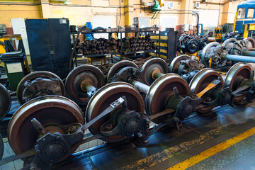 Train wheels and chassis are organized neatly in a railway workshop. Workers move around, busy with repairs and maintenance tasks in the bright space filled with tools and machinery