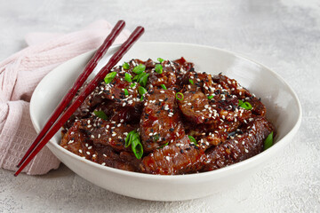 Crispy beef, with green onions and sesame seeds, in a bowl with chopsticks, on a textured surface, on a light background, no people