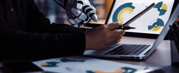 A close-up view of a human hand collaborating with a robotic hand over a laptop, analyzing data and charts in a modern office setting, showcasing advanced technology.Noogenesis