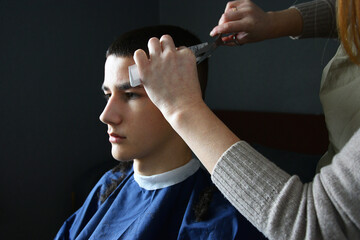Close up of hairdresser trimming bangs of teenage boy with scissors and white comb in salon