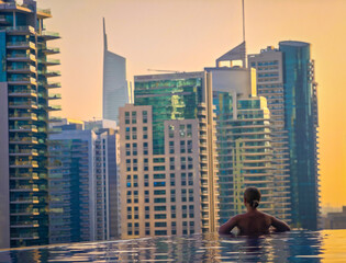 Woman relaxing in infinity pool at sunset, watching city skyscrapers, luxury lifestyle and urban skyline view. Dubai background
