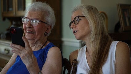 Elderly senior woman and her daughter look at pictures and enjoy video calls while sitting together indoors in a home setting