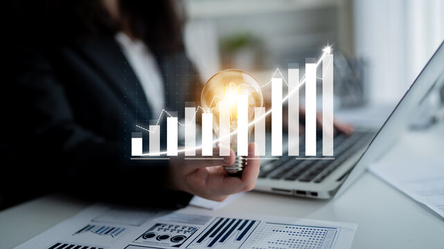 A business professional holds a light bulb while analyzing financial data on a laptop. The image conveys themes of growth, innovation, and strategic planning in an office setting. Impute - Powered by Adobe