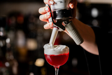 A bartender is adding a creamy white foam topping to a vibrant red cocktail served in an elegant stemmed glass at a bar with a dark, moody atmosphere