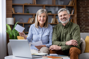 Happy senior couple sitting on sofa at home, smiling while using a laptop and reviewing documents...